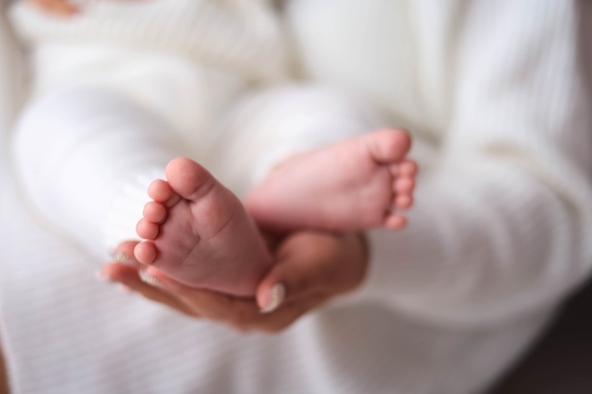 白いニットにくるまれた赤ちゃんの足を、親が両手でそっと包んでいるところ
