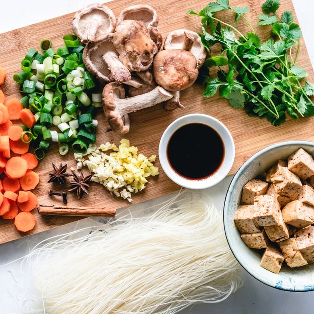 まな板の上に並べた豆腐・しいたけ・青ねぎ・にんじん・香菜と、米麺と醤油。食材を分散させた明るい俯瞰の食材フラットレイ