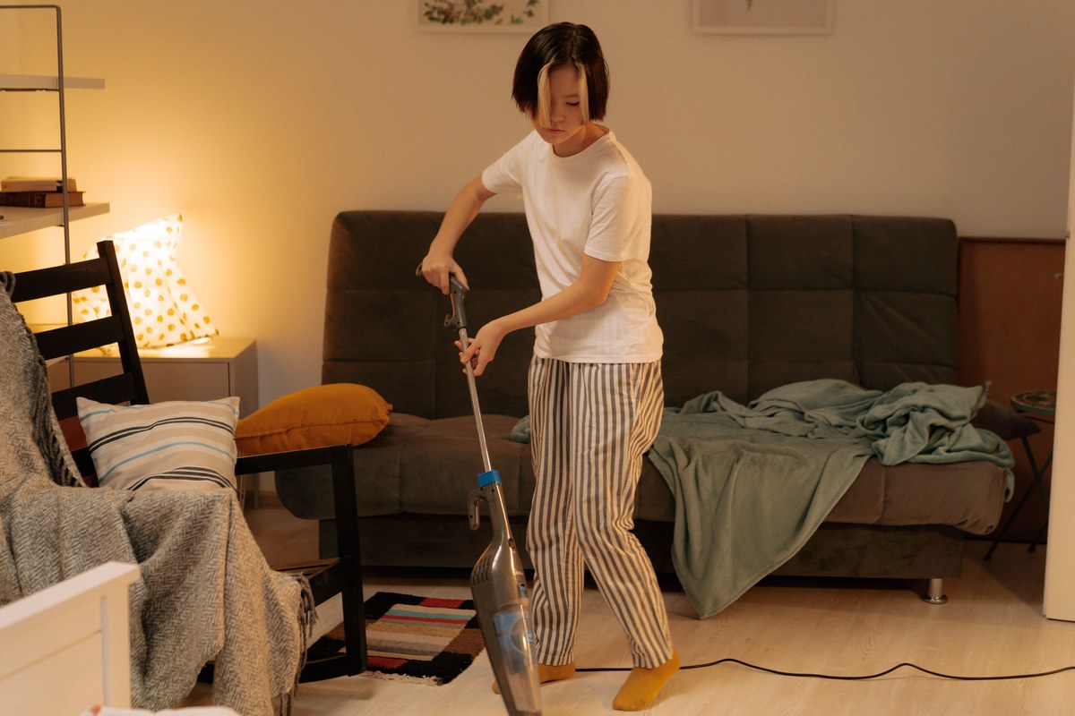 寝室でスティック型の掃除機をかけている女性。奥のベッドには寝具が広げられている
