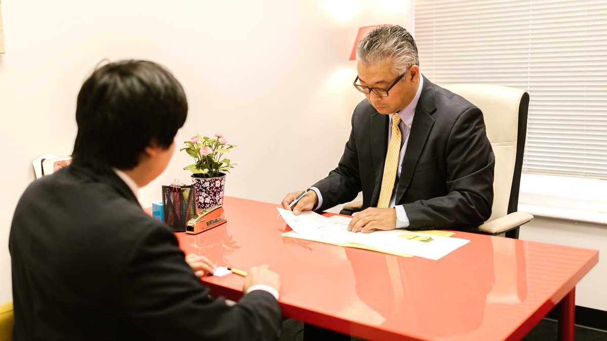 明るい室内で机をはさみ、書類を見ながら落ち着いて話す東アジア系の男性2人