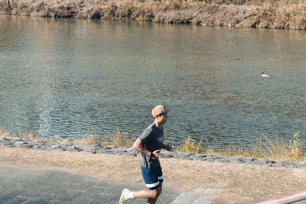 川沿いの遊歩道をジョギングしている男性。ランニングキャップと半袖姿で、背景に川と対岸の堤防が写っている