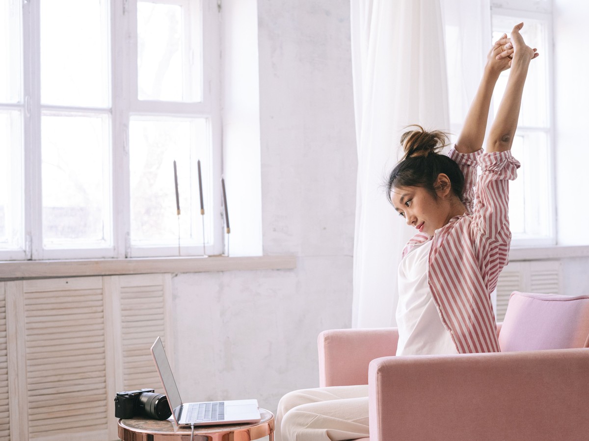 明るい部屋で、ノートパソコンから離れて両腕を上に伸ばし、ひと休みする女性