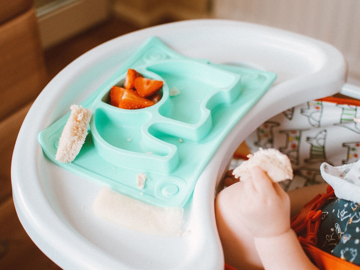 하이체어 식판 위의 이유식과 빵 조각을 쥔 아기 손, 곁에 턱받이가 보이는 클로즈업
