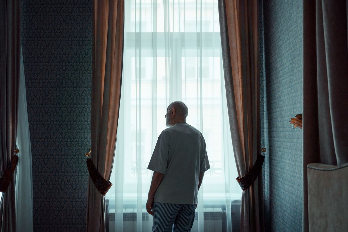 An older man stands quietly at a bright window, seen from behind, gazing outside.