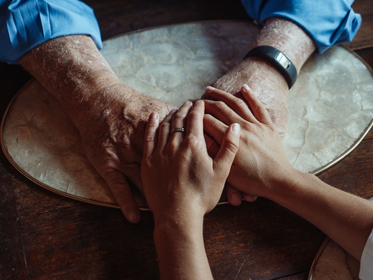 A younger person's hand rests gently over an older parent's hands in warm light.