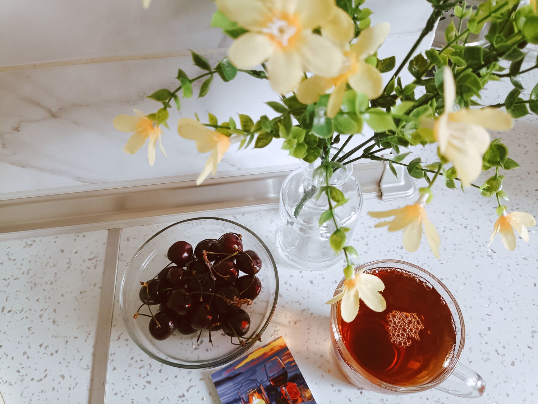 A calm, bright flat-lay on a light table: a warm cup of tea, a glass bowl of fresh cherries, and a small vase of yellow flowers