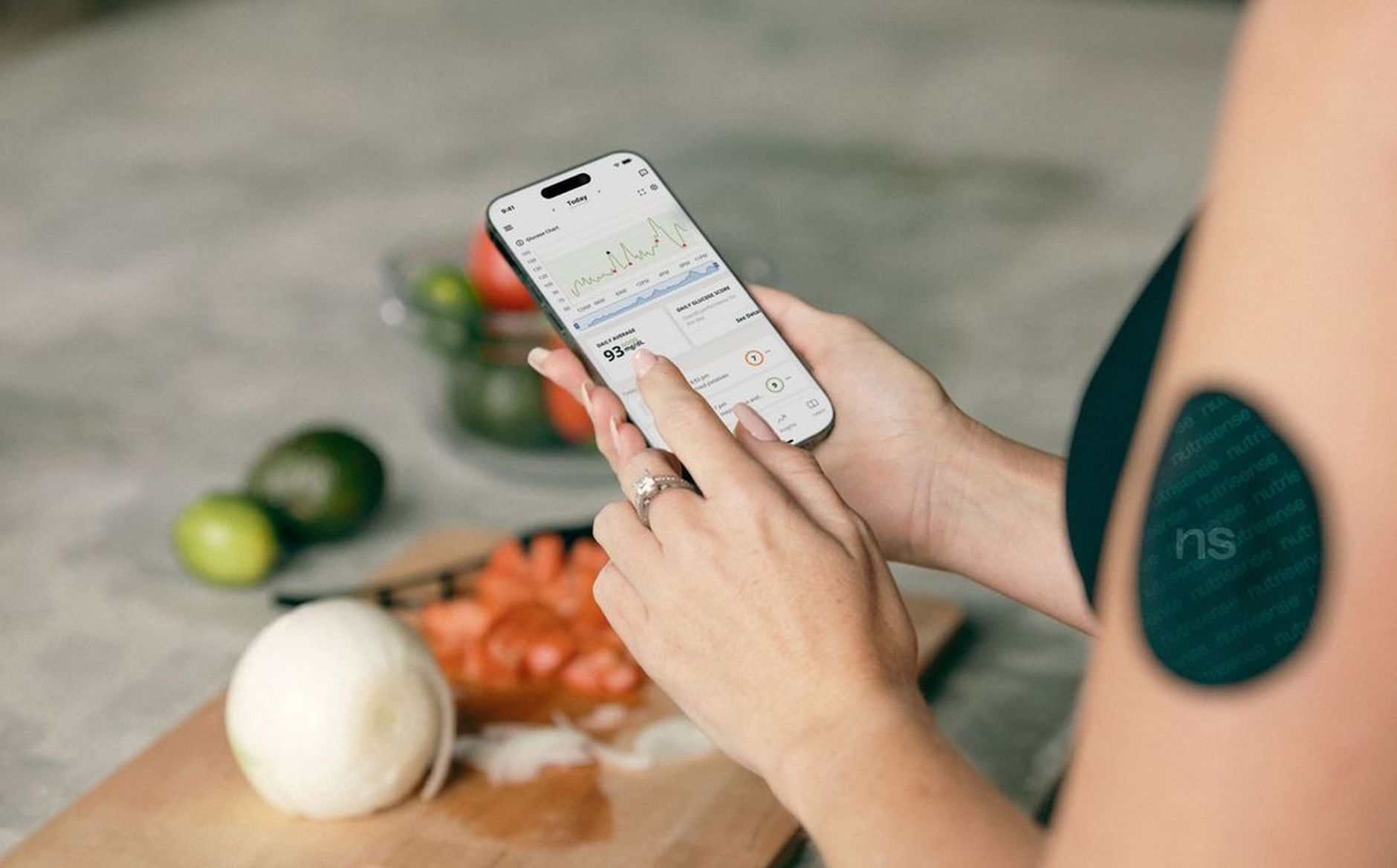 A CGM sensor on the back of the arm with a smartphone showing a glucose trend chart in a kitchen