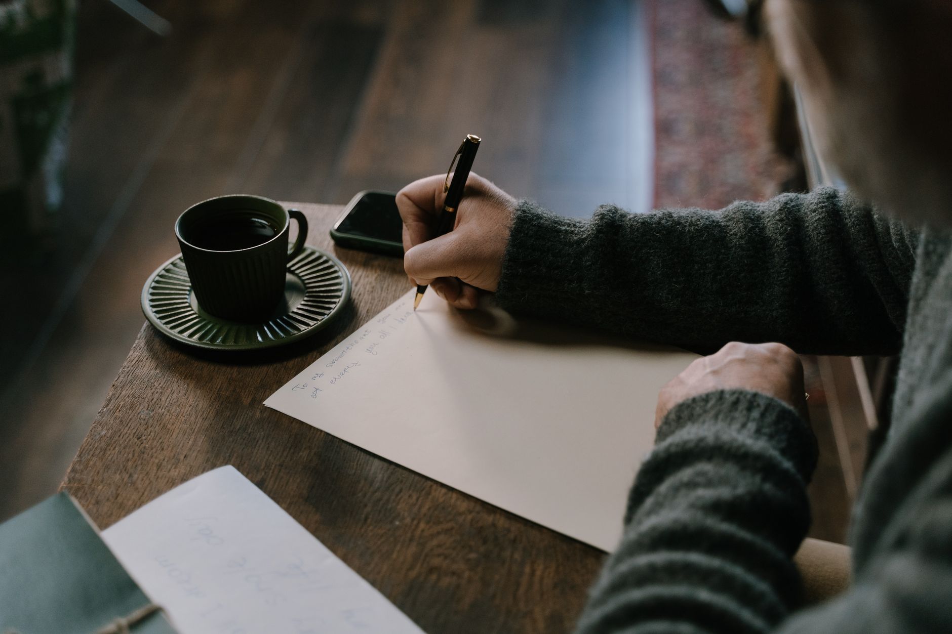 A person writing by hand in a notebook beside a cup of coffee in warm natural light