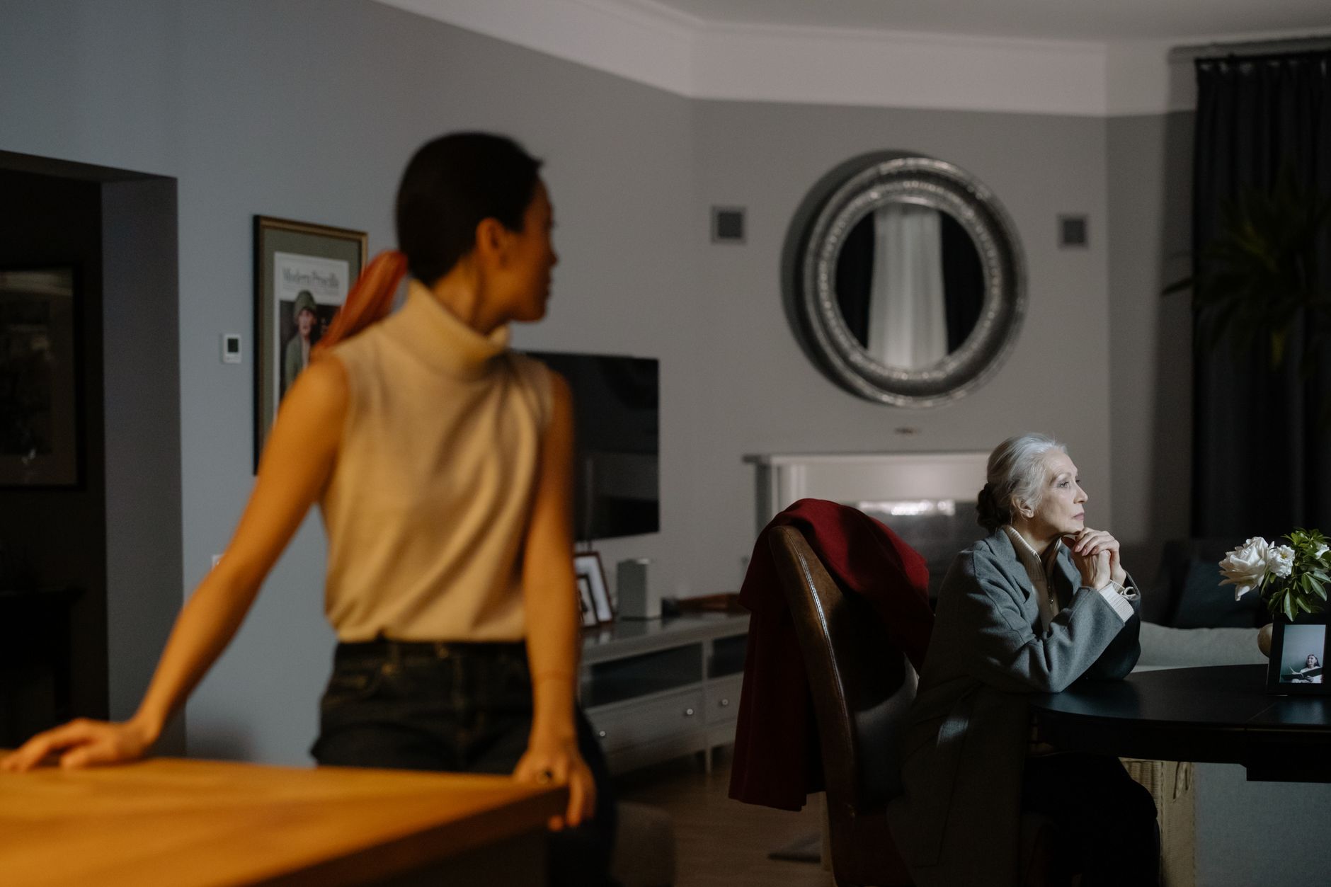An older woman sitting quietly and withdrawn at home while a younger family member stands apart