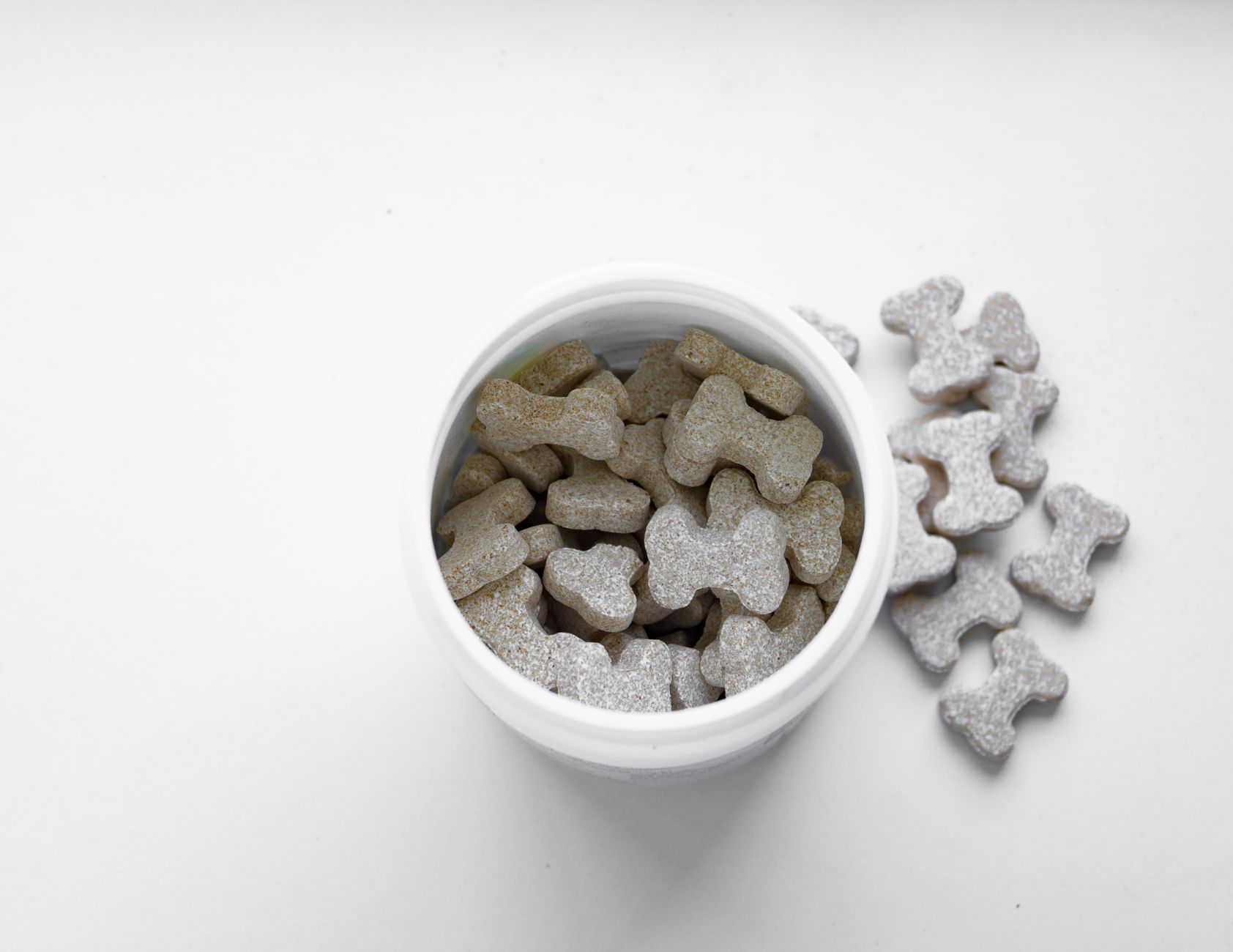 Bone-shaped dog dental treats in a white container on a clean white surface, an adjunct to brushing rather than a substitute