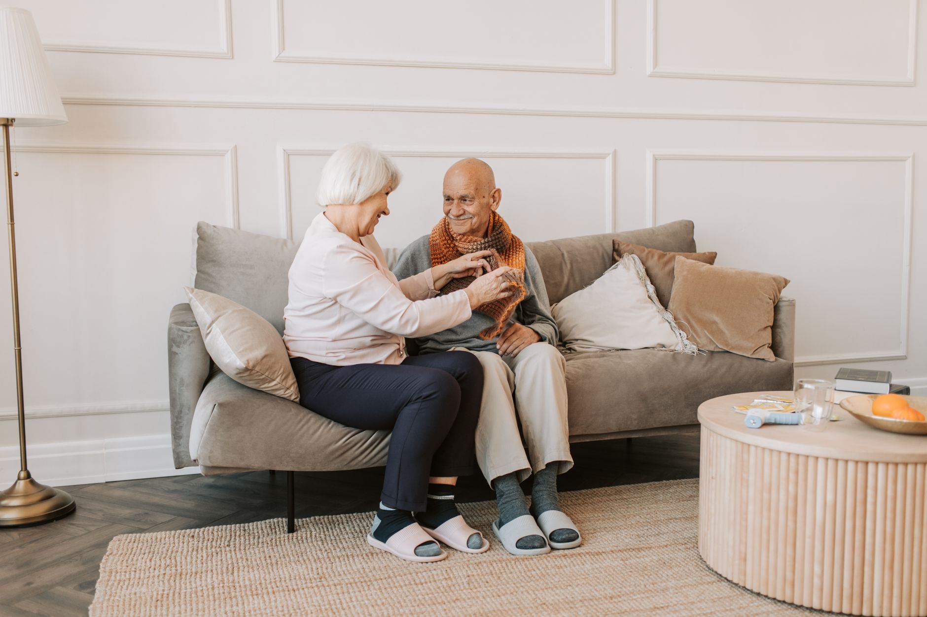 An older couple sitting together at home, one gently adjusting the other's scarf — everyday comfort and care