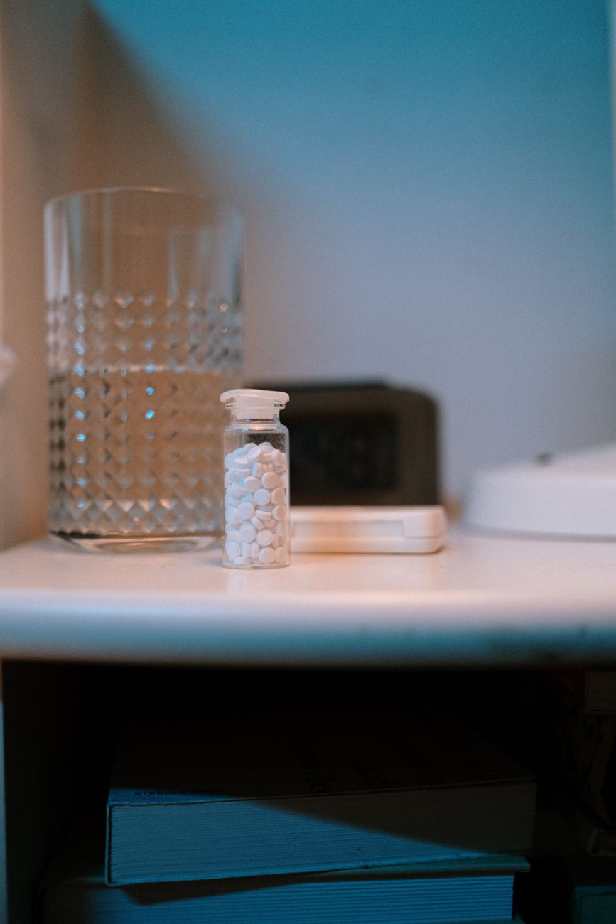 A glass of water and a bottle of tablets on a nightstand in a calm, dimly lit bedroom
