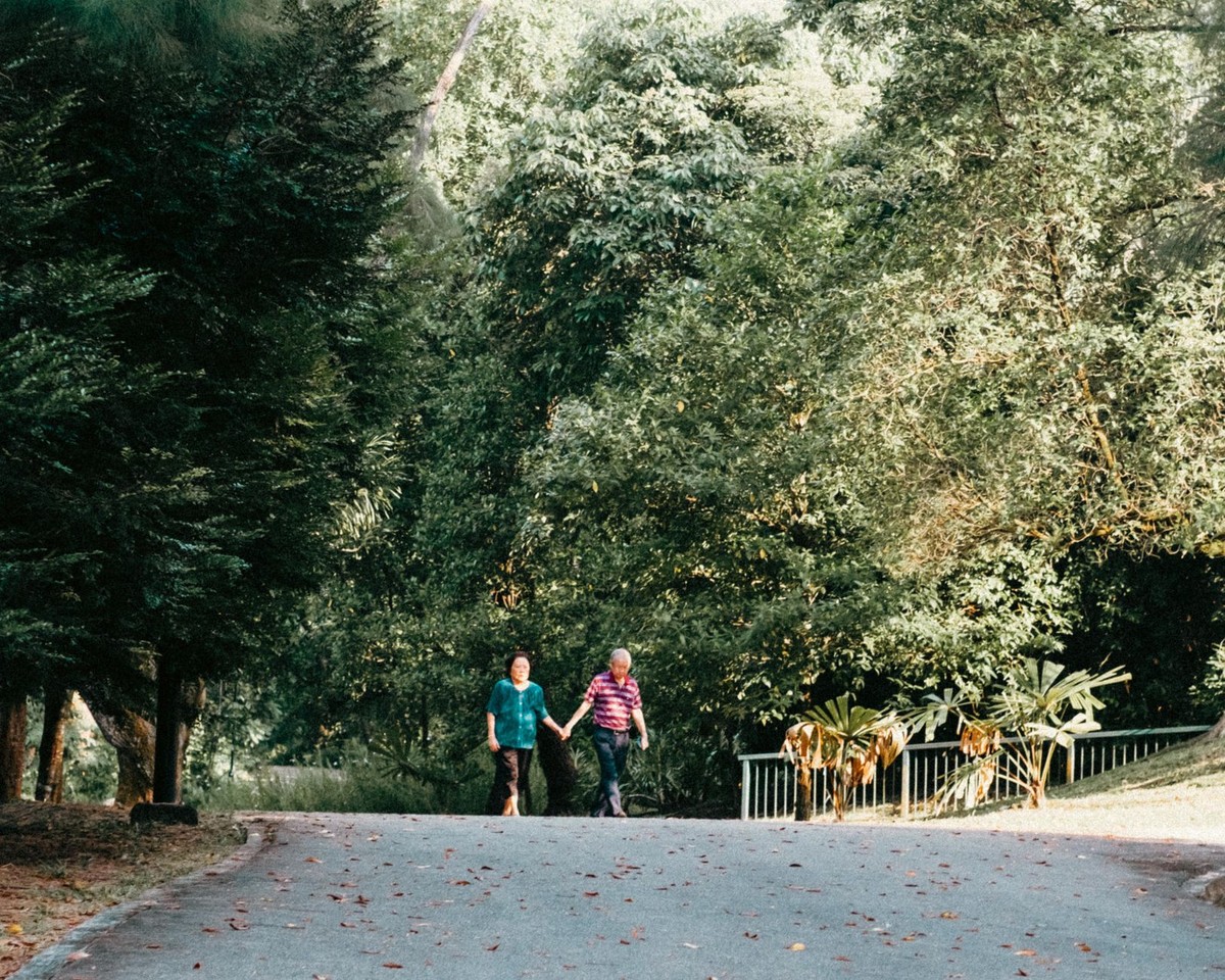An older adult and a younger family member walk together along a green, tree-lined path.