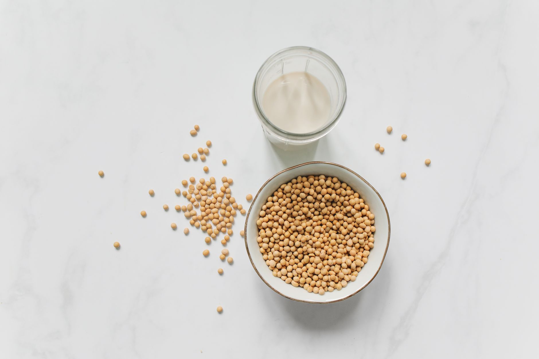 A bowl of soybeans and a glass of soy milk on a white marble surface