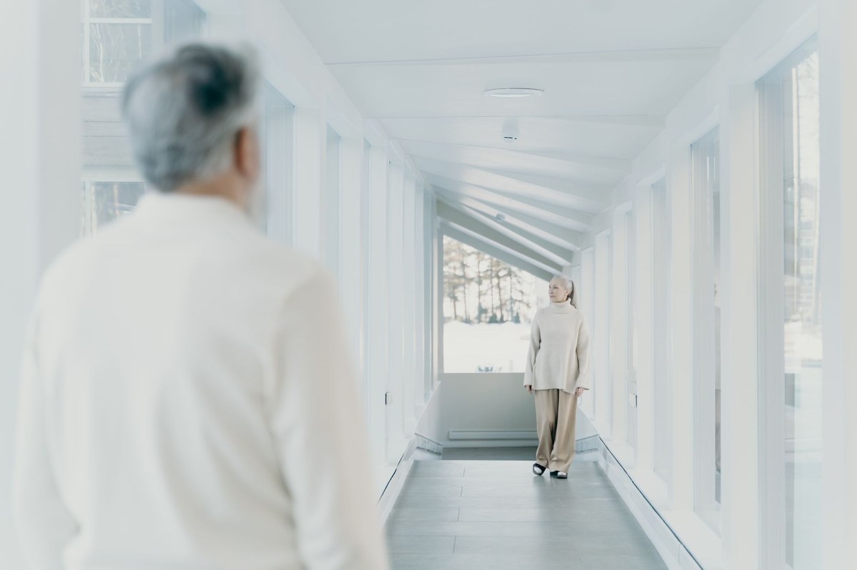 A senior woman walking heel-to-toe for balance practice down a bright, window-lined hallway