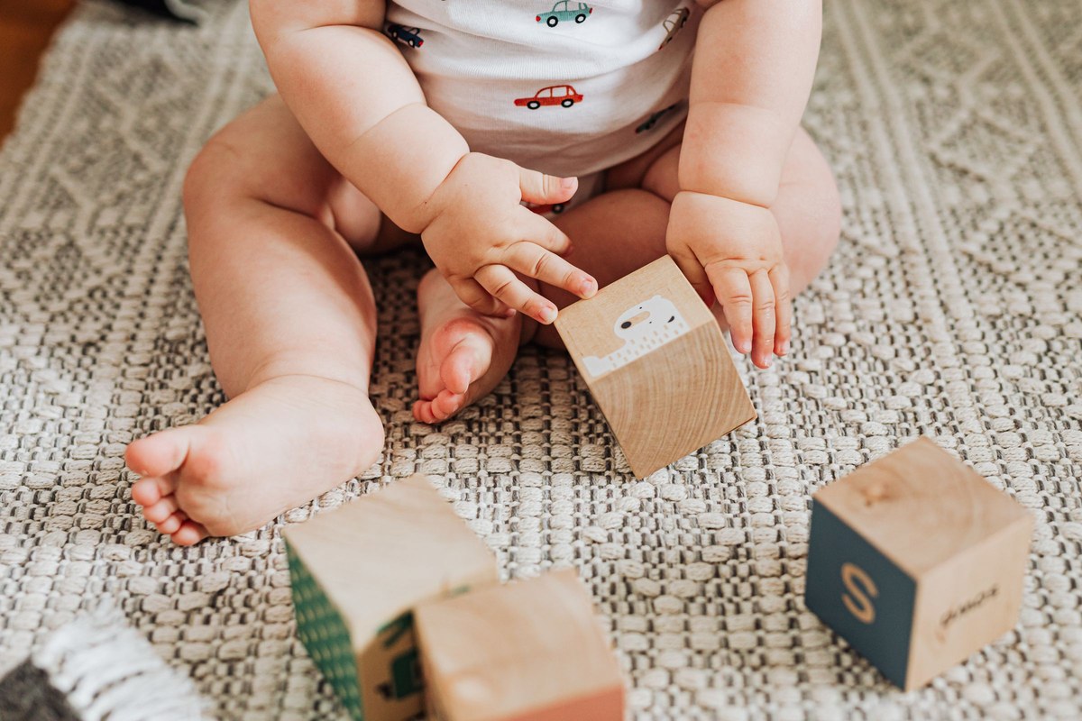 A baby sitting on a woven rug turning a wooden block over in both hands, the ordinary play a dehydrated baby stops doing