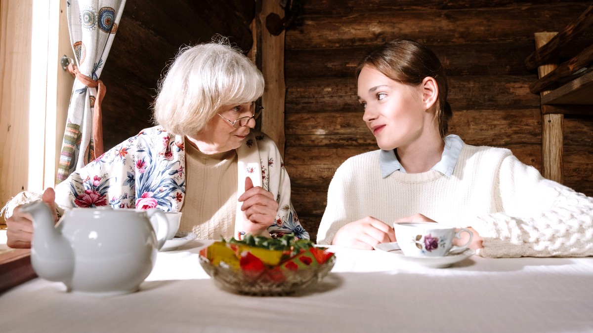 An older woman and a younger woman sitting across a cloth-covered table with a teapot, cups and a bowl of food between them, talking in the light from a nearby window