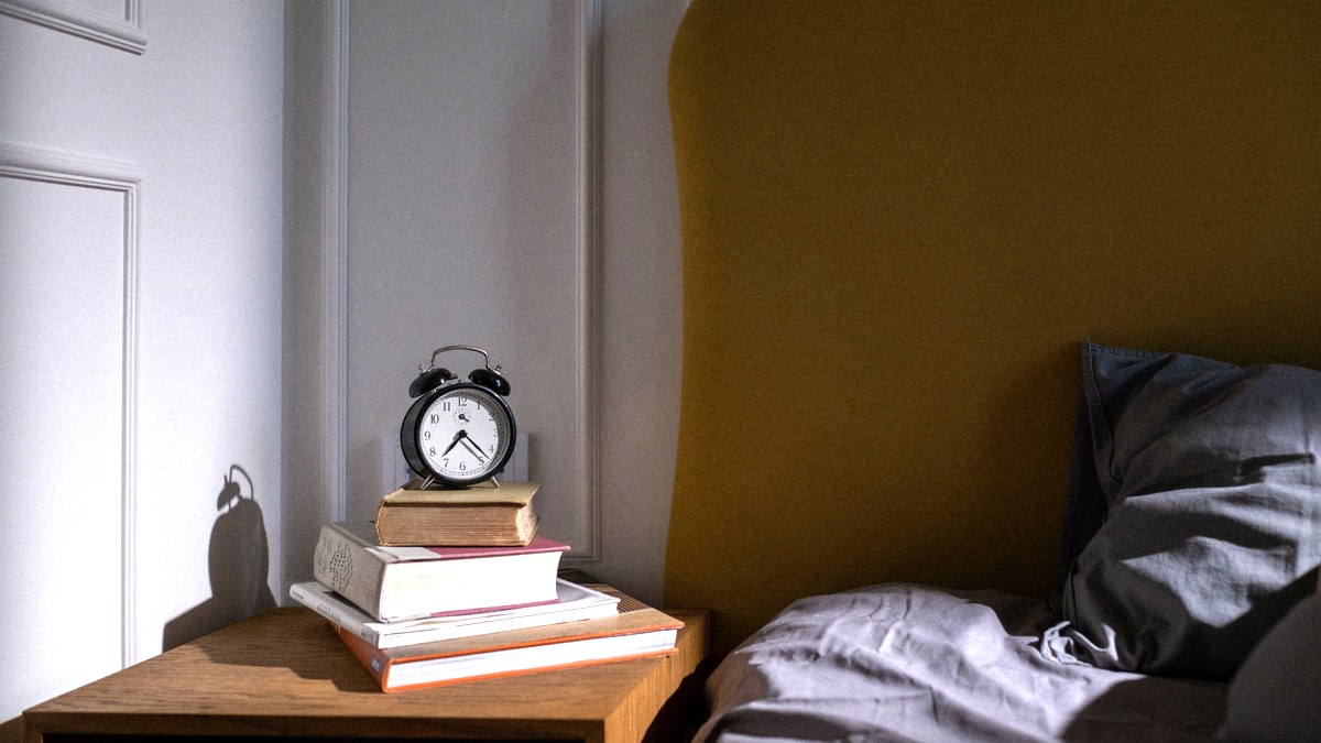 A quiet bedroom corner at first light, an alarm clock and a stack of books on the nightstand beside an empty bed