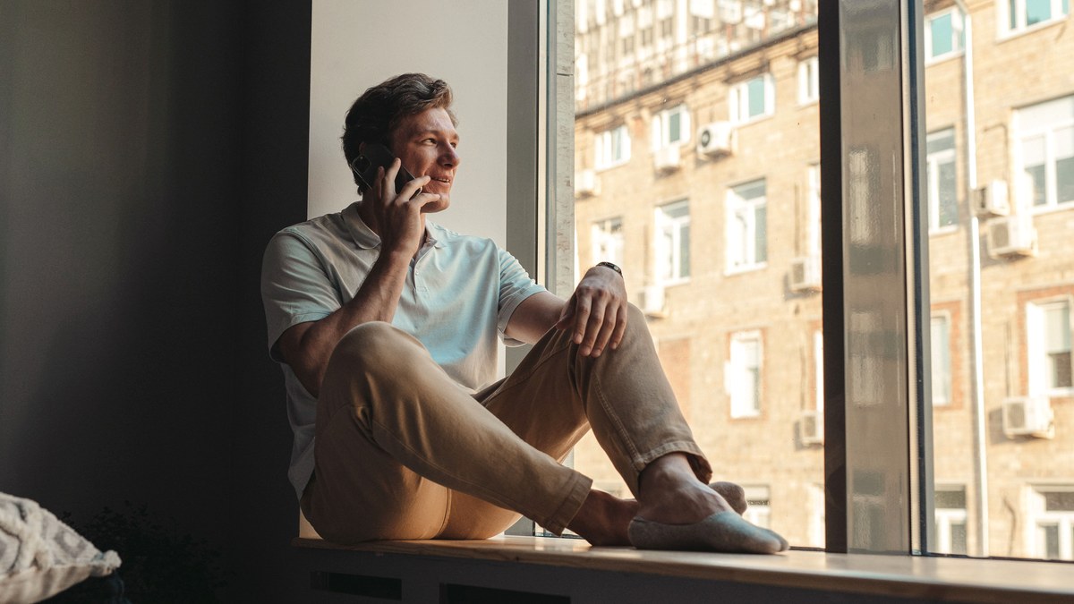 A man sitting at a large window in morning light, mid-conversation on a phone call, seen from the side