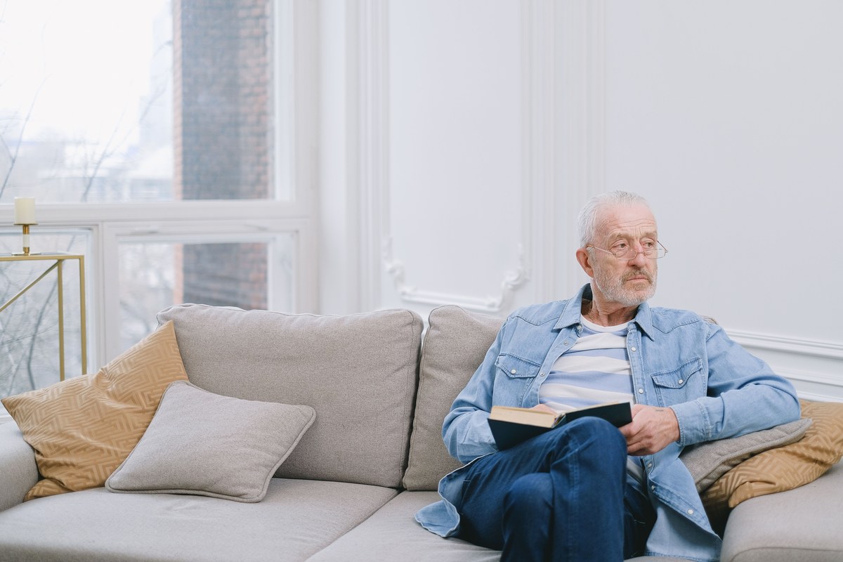 An older man sitting with an open book on a pale sofa in a bright, window-lit living room, pausing to look up