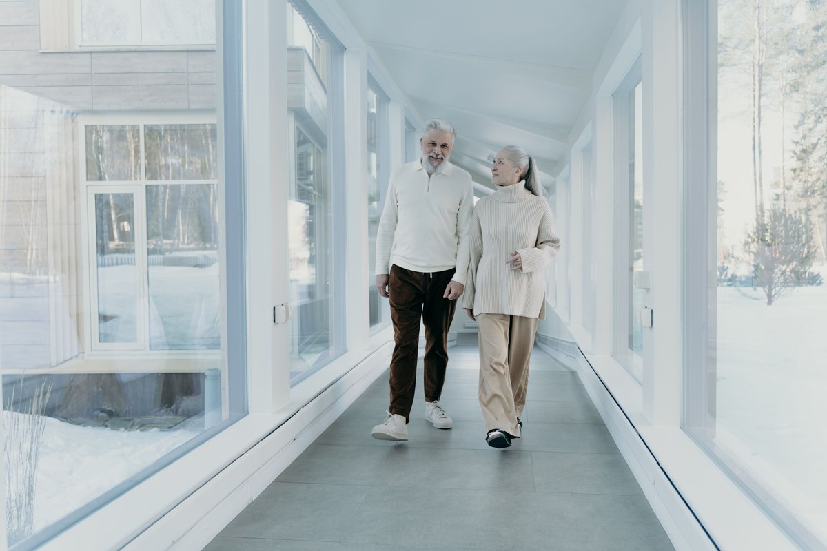 An older couple walking steadily side by side down a bright glass-walled corridor in daylight