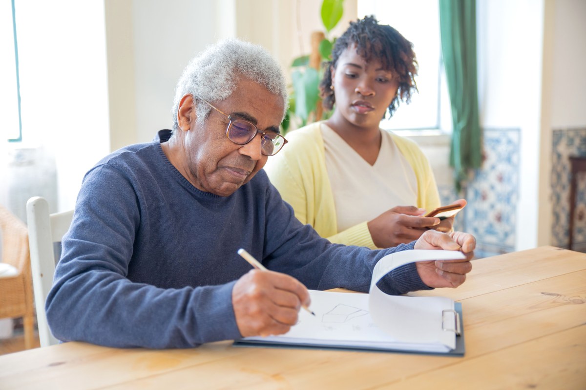 An older man in glasses writing on a clipboard at a sunlit wooden table while a younger woman sitting beside him reads from a paper list.