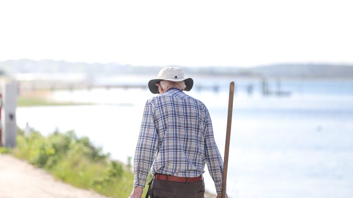 An older man in a sun hat walking away along a bright coastal gravel path with a wooden walking stick in one hand.