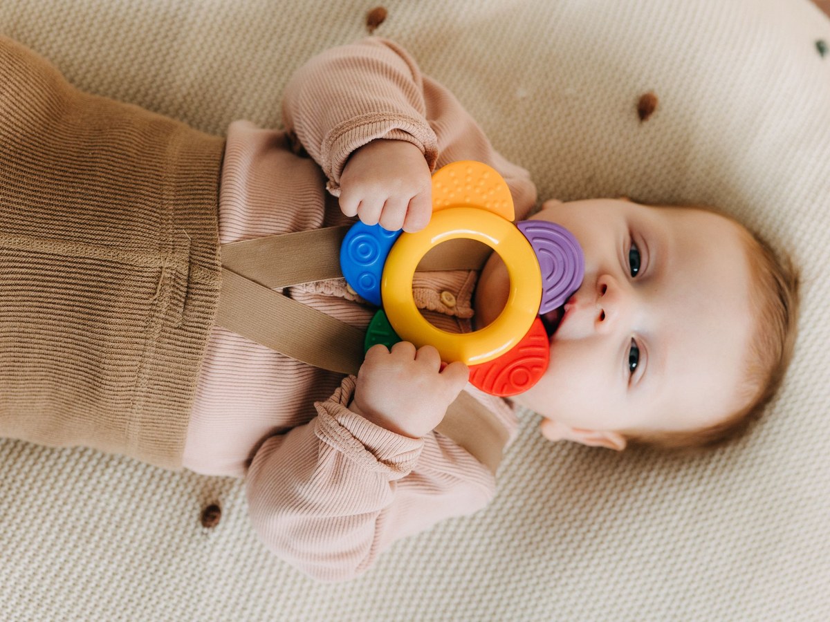 A baby lying on a knit blanket chewing on a colorful silicone teething ring held in both hands