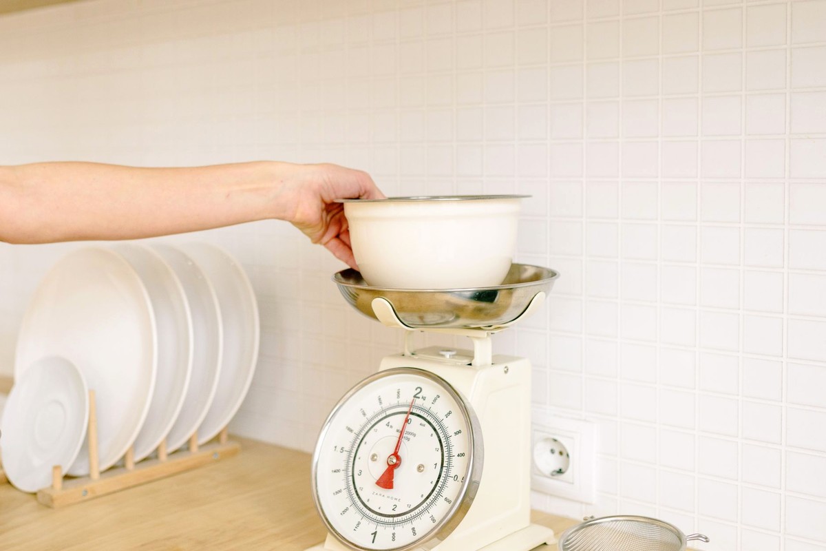A hand setting a white bowl onto a kitchen weighing scale on a bright, tiled counter