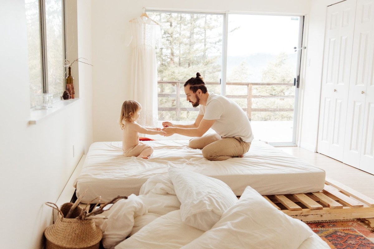 A father and his toddler sitting together on a low bed in a bright, sunlit bedroom, gently holding hands