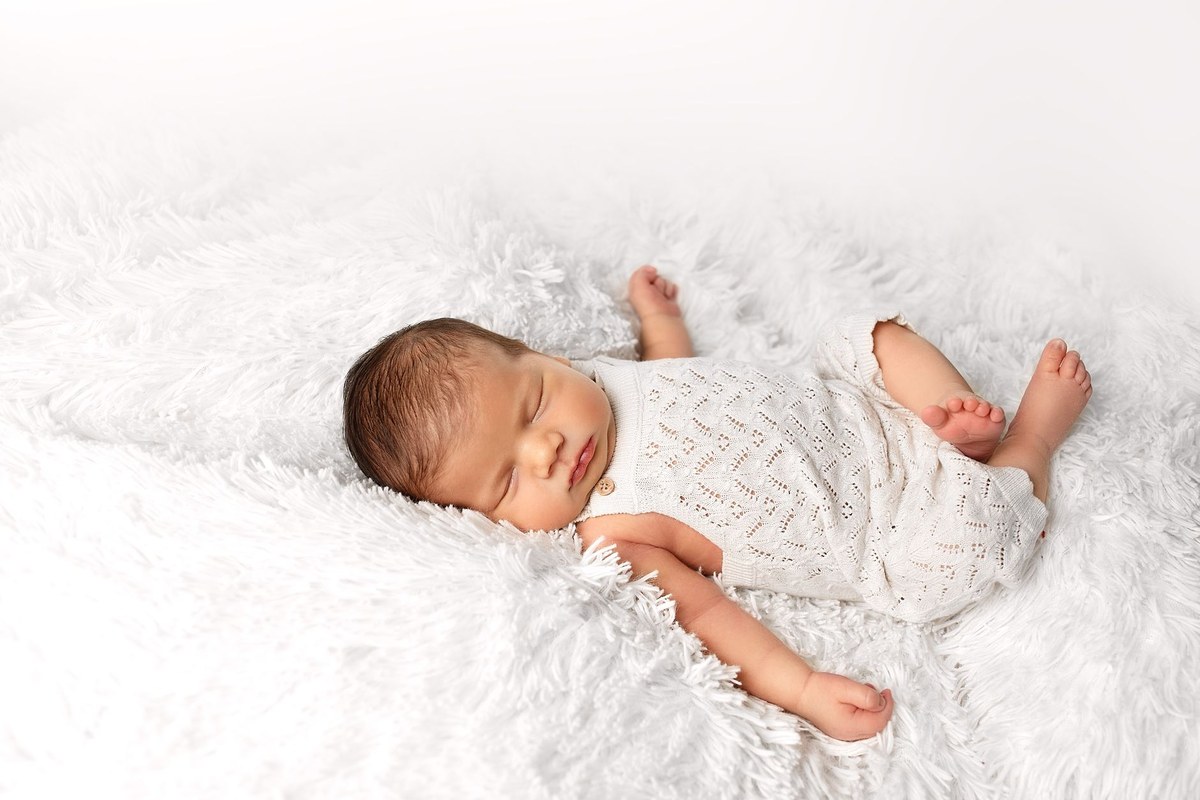 A newborn in a white knit romper sleeping peacefully on a soft white blanket