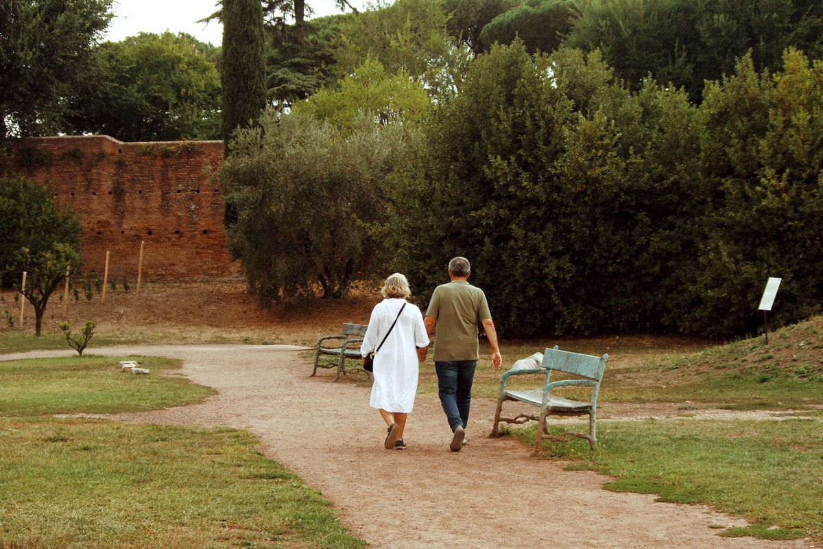 An older couple walking away from the camera along a gravel path through a green park, wooden benches and trees on either side