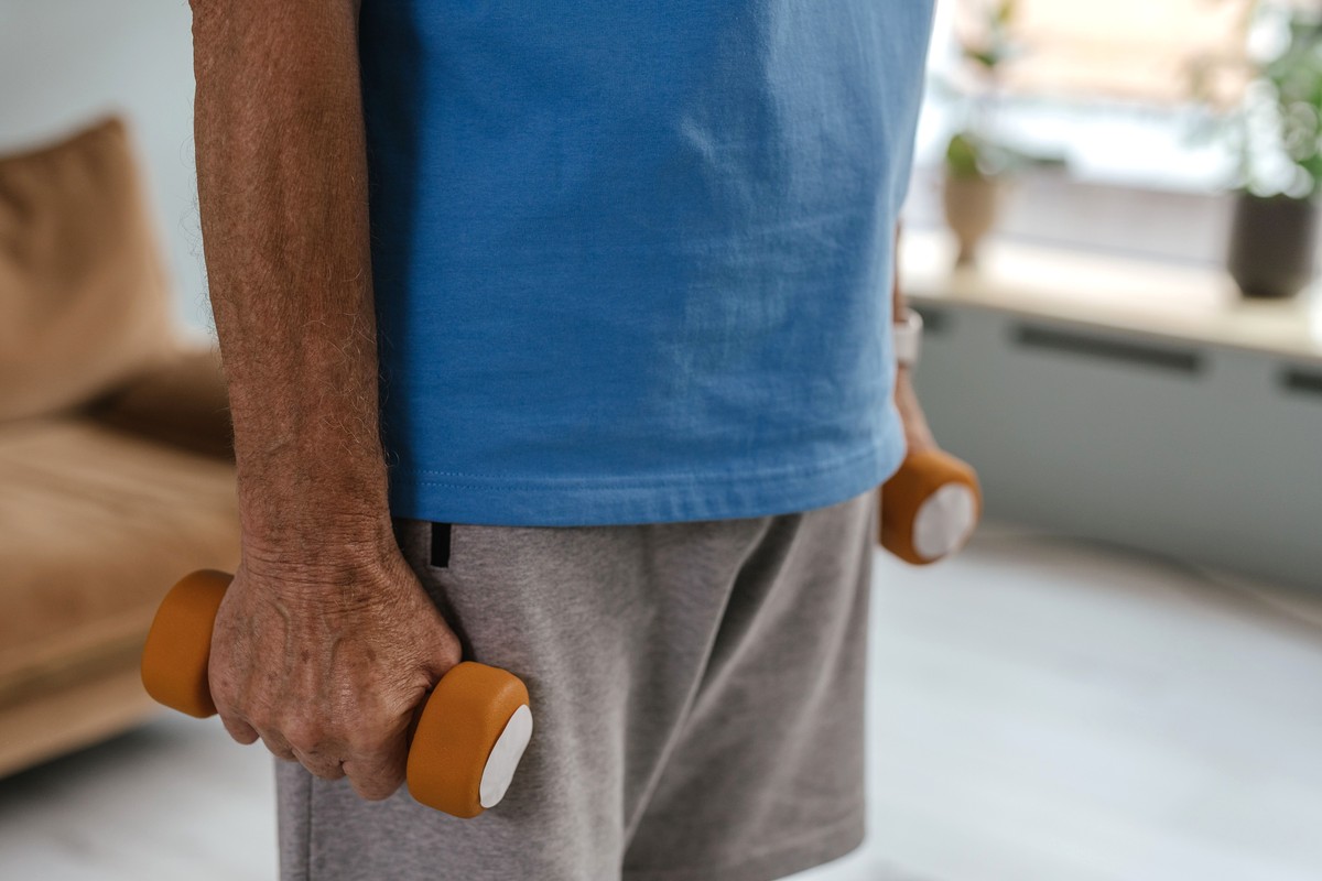 An older person standing in a bright living room holding a pair of small orange dumbbells at their sides, sofa and window plants behind them, face not shown