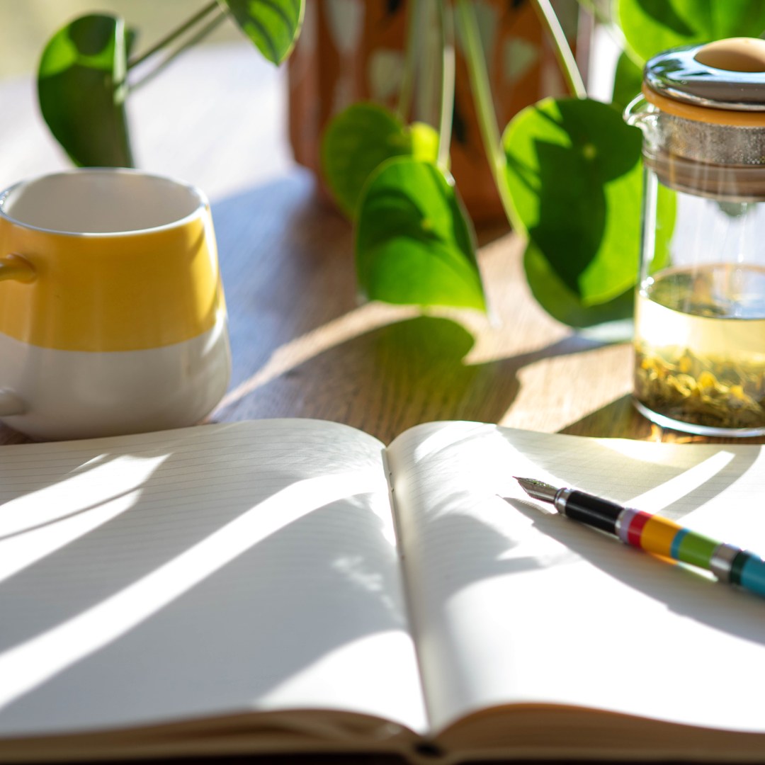 An open blank notebook and a striped fountain pen on a sunlit wooden table beside a yellow mug, a glass tea infuser and a green houseplant.