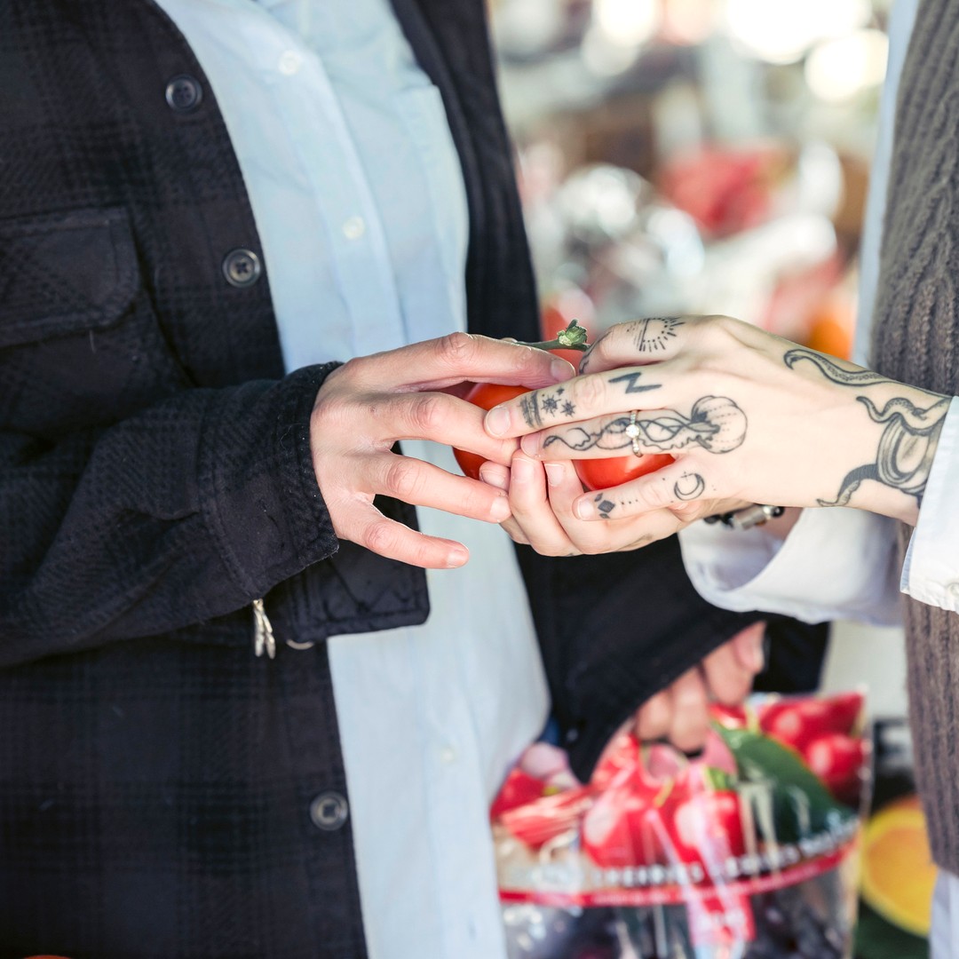 Two pairs of hands examining a ripe red tomato together at an open-air produce market stall, crates of fruit and vegetables blurred behind them