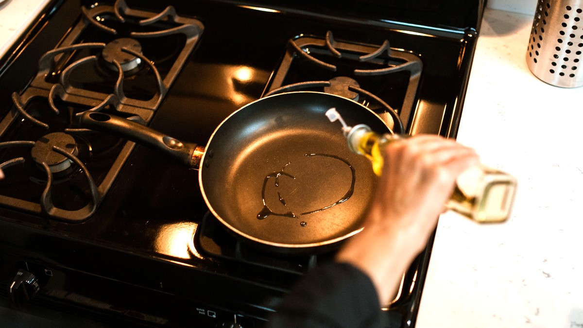 A hand pouring olive oil from a bottle into a frying pan on a gas stove, seen from above