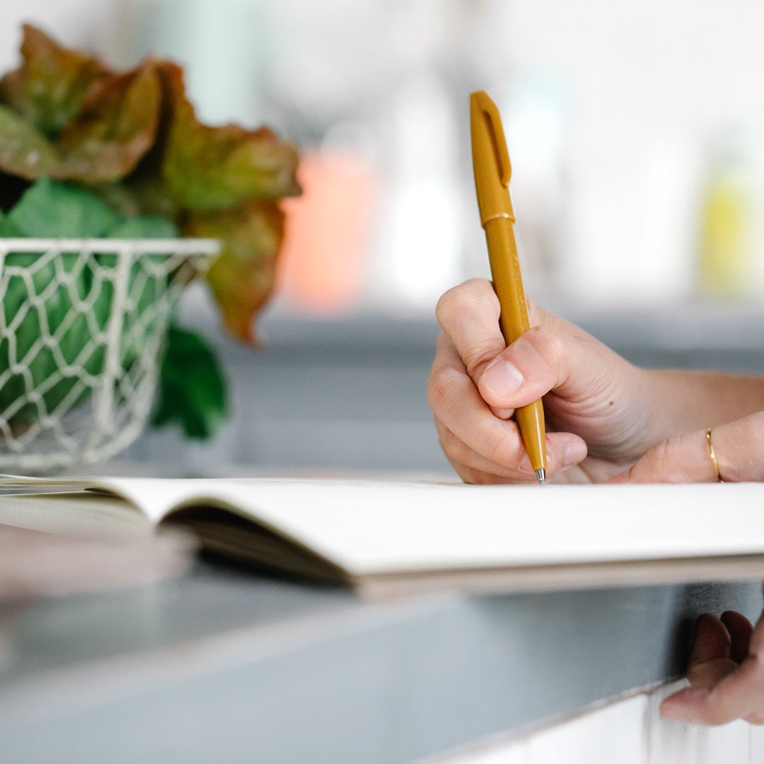 A hand writing in an open paper notebook on a kitchen counter beside a wire basket of fresh greens, in daylight
