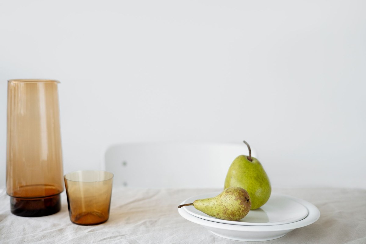 Two ripe green pears on a white plate beside a small amber glass and carafe on a linen-covered table in soft daylight