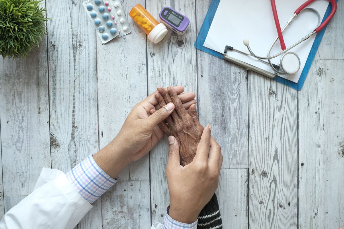 Overhead view of a clinician's hands holding an older patient's hand on a desk laid with pill bottles, blister packs, a clipboard and a stethoscope