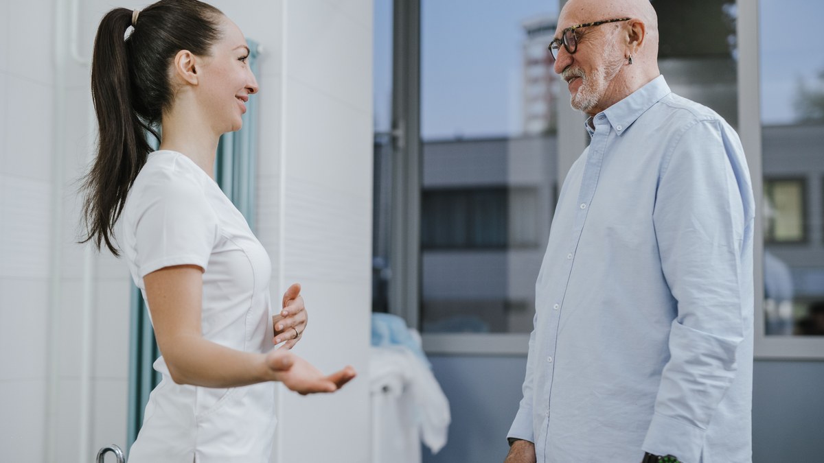 Older man in a light blue shirt talking with a nurse in a bright clinic corridor