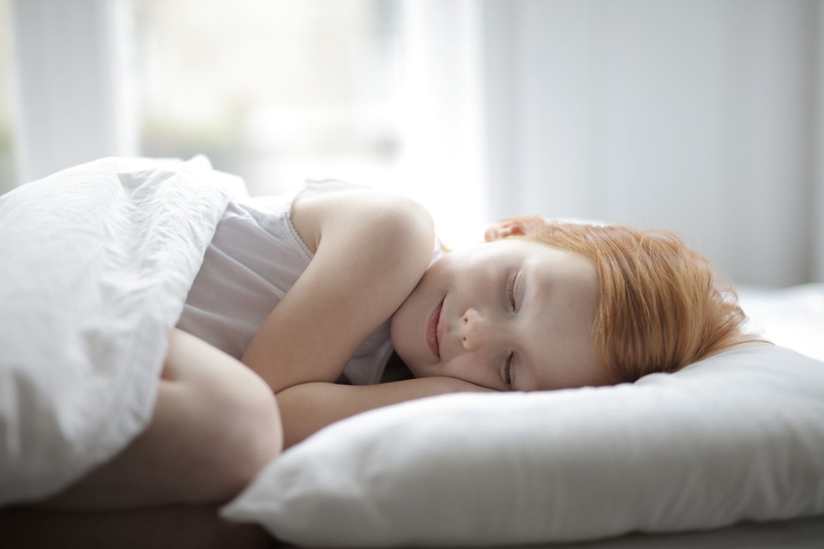 A young child asleep on a white pillow under light cotton bedding in a bright, airy bedroom