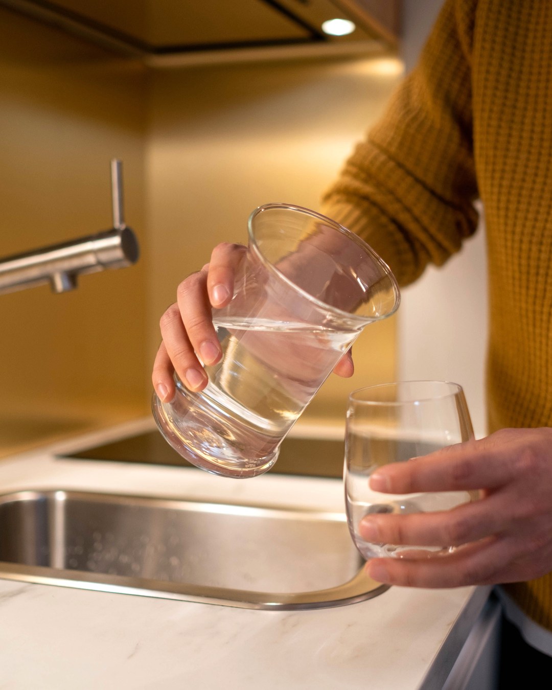 A hand pouring water from one glass into another at a kitchen counter