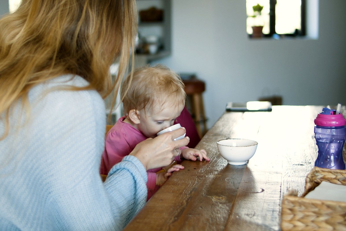 A parent holding a small cup to a young child's mouth at a kitchen table, giving a few sips at a time