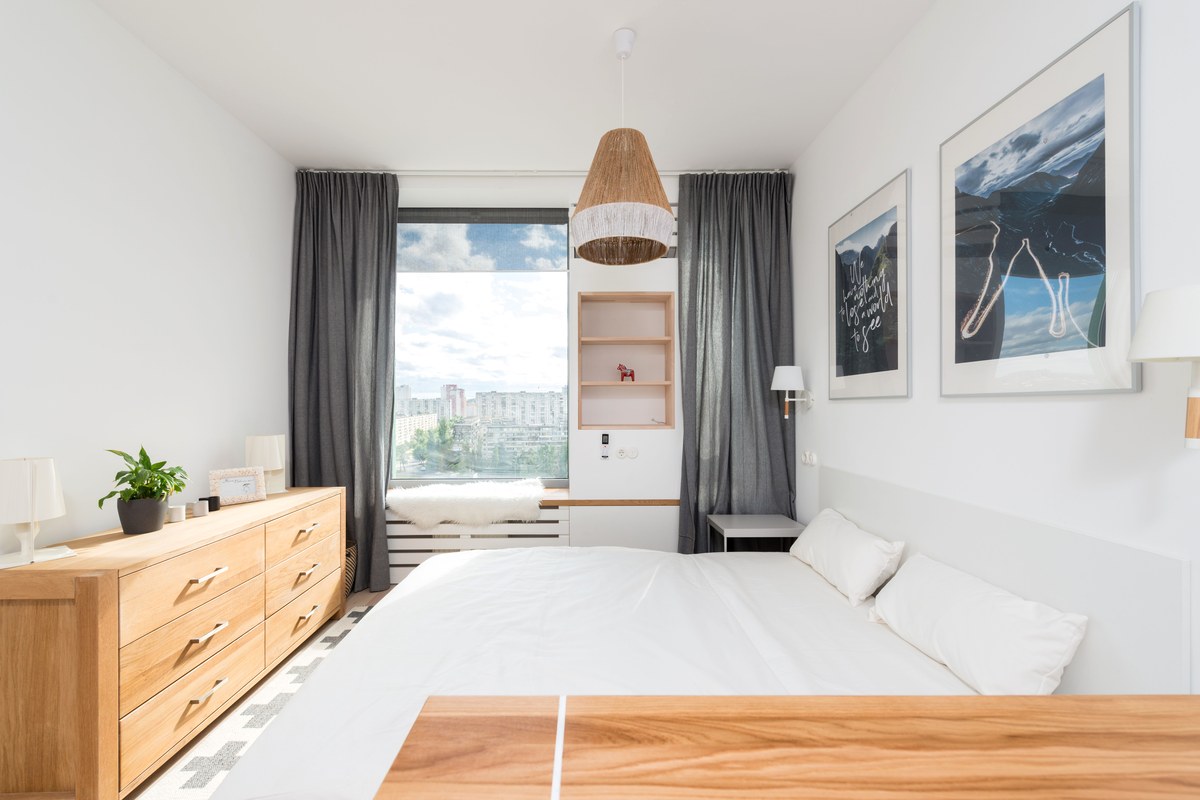A neatly made white bed in a bright, airy bedroom with the curtains drawn open to a daylit window.