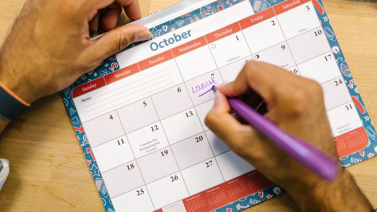 A hand marking a date on a wall calendar with a purple pen on a wooden desk