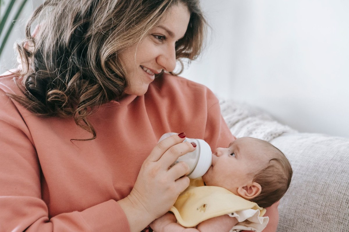 A caregiver bottle-feeds a baby cradled upright in the crook of the arm in soft natural light