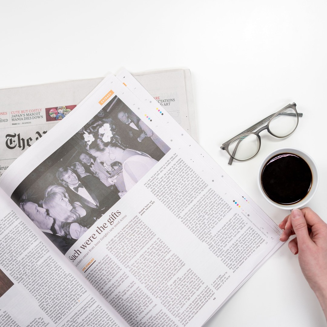 Overhead view of an open newspaper on a white table with reading glasses and a cup of black coffee beside it