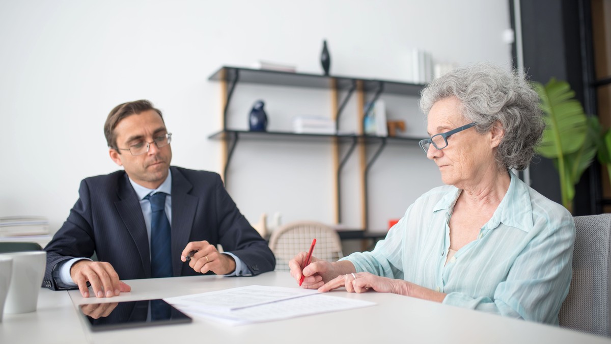 An older woman with gray hair and a professional in a suit seated at a white table in a bright room, talking calmly over a printed sheet of paper