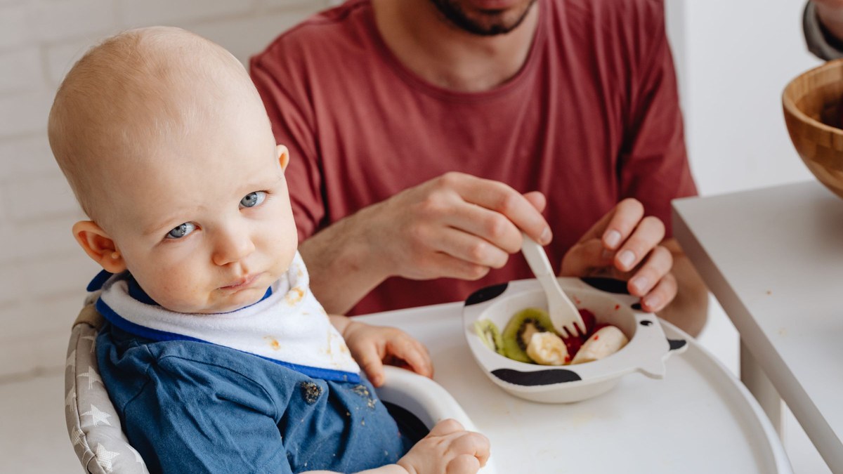 A parent offering a baby in a high chair a bowl of soft fruit in a bright white kitchen