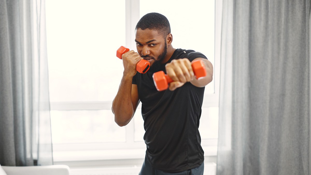 A man in a t-shirt doing a simple dumbbell exercise beside a bright window at home